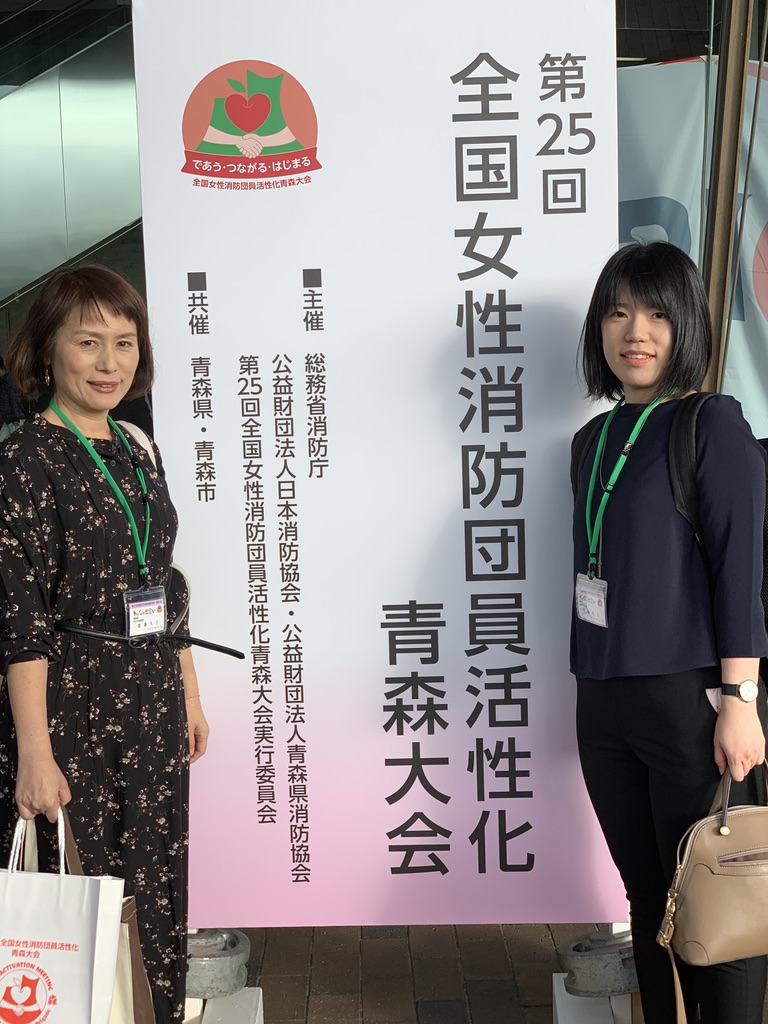 第25回全国女性消防団員活性化青森大会の看板の両脇に参加したカメリア隊の女性2名が並んで記念撮影をしている写真