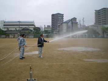男性一人がホースを支え、もう一人が散水ノズルを持って散水を行っている様子の写真