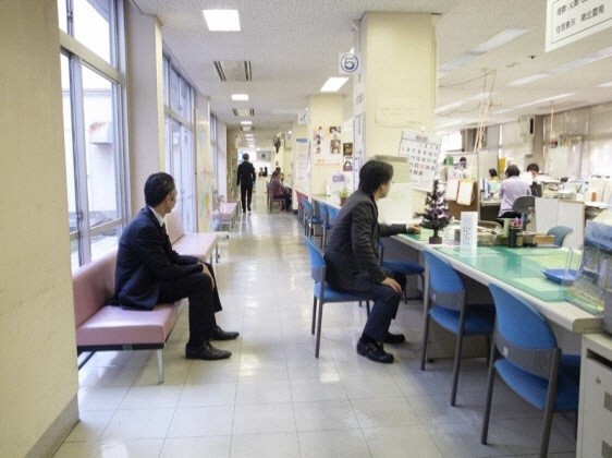 窓口とガラス窓の間の狭い通路に設置されたピンク色の長椅子と机に設置された青い椅子に来庁者が座っている写真