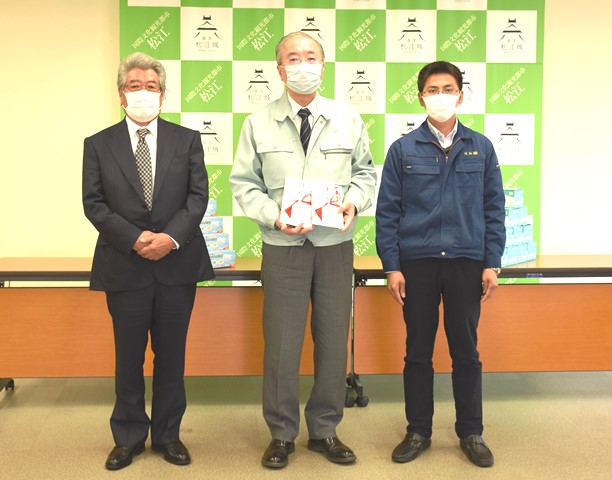 テーブルに並べられたマスクを背景に、目録を2つ持っている松浦松江市長と寄贈者の男性2人の写真