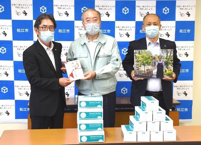テーブルに並べられたマスクを前に、松浦松江市長と一緒に目録を持っている男性、張様の写真を持っている男性の写真