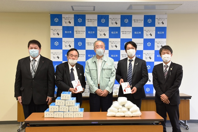 テーブルに並べられたマスクを前に、松浦松江市長を挟み目録を持って笑っている寄贈者の方々の写真