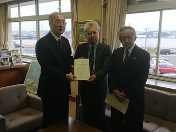スーツを着た三人の男性が机とソファの間に並んで立ち、左の上座の前に立つ男性と中央の男性が一緒に書類を持ち、報告書提出の様子を写した写真