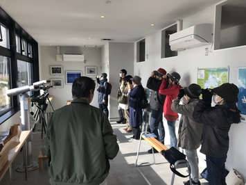 室内で9人の男女が双眼鏡を覗き鳥を観察している写真