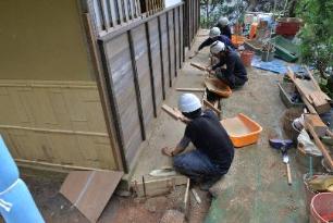 御風呂屋の西側の外壁に沿って土間叩きを施工している4人の作業員の写真