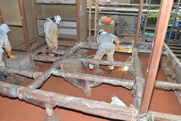 格子状に組まれた木材の隙間に立って床下土壌の鋤取り作業を行っている3人の作業員の写真