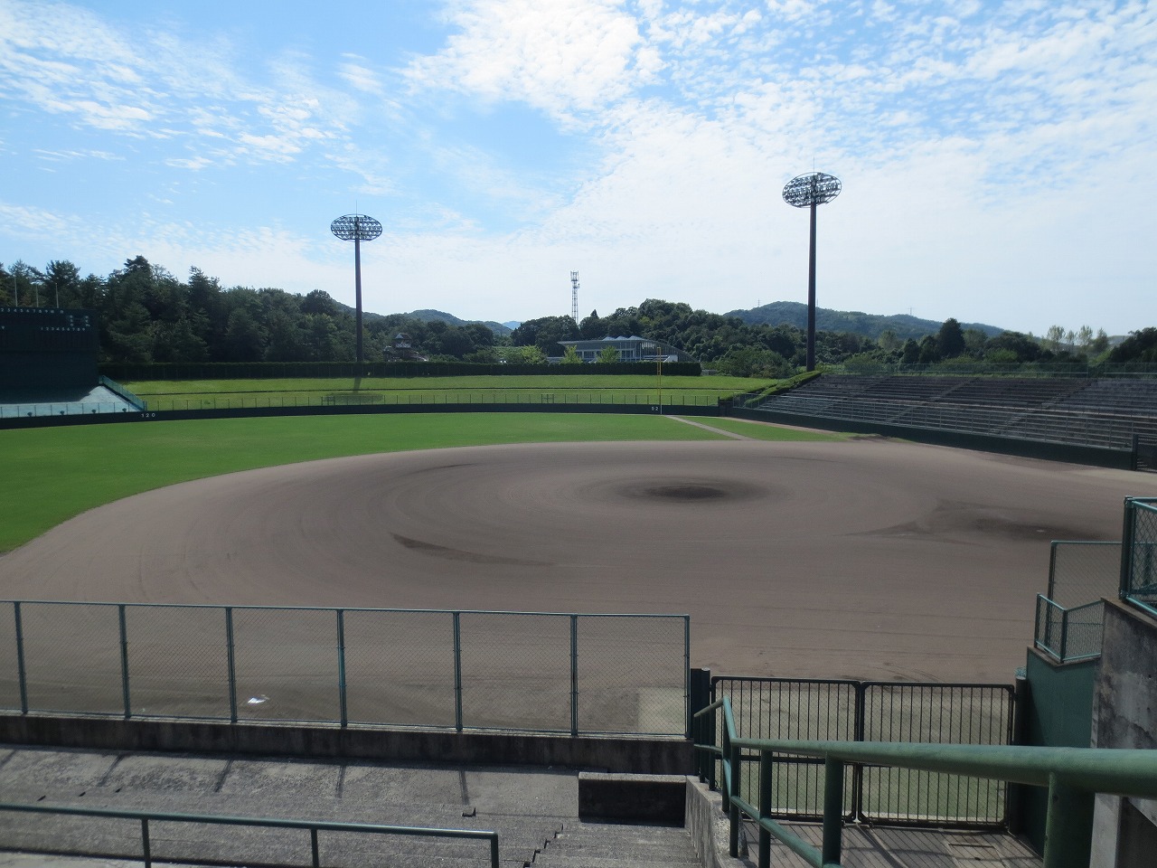 白い雲と青い空を背景に、土でできた内野とその周囲を芝生で覆われた外野で構成された野球場の写真