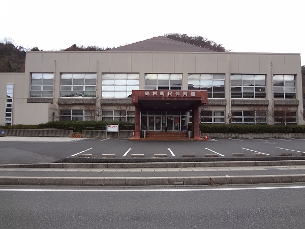 くもり空の下、砂色の壁と茶色のエントランスの島根町民体育館の正面からの写真、建物の前には12箇所の駐車場が見えている