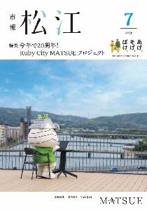 7月号表紙。市役所テラスでパソコンを使って仕事をしているおまっちぇの写真です。