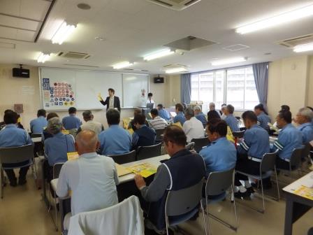 指導員研修でホワイトボードを使い説明をしている人の話を座って聞きながら資料を見ている指導員の方々を部屋の後方から写した写真