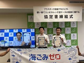 使い捨てコンタクトレンズ空ケースの回収に関する協定式で男性二人とモニター越しの男性が締結書を持っている写真