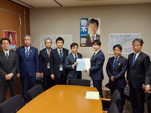 8名の男性が横一列に並び、中央に立つ青木一彦参議院議員と同盟会の男性が一緒に要望書を持っている集合写真