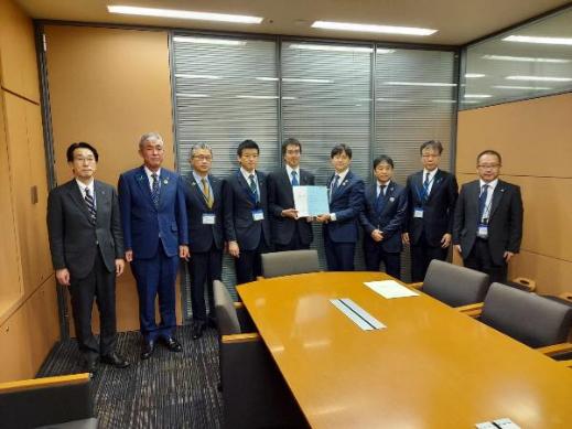 9名の男性が横一列に並び、中央に立つ高見康裕参議院議員と同盟会の男性が一緒に要望書を持っている集合写真