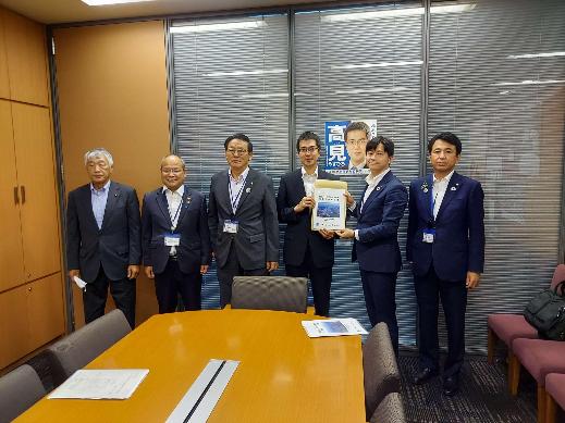 6名の男性が横一列に並び、中央に立つ高見康裕衆議院議員と同盟会の男性が一緒に要望書を持っている集合写真