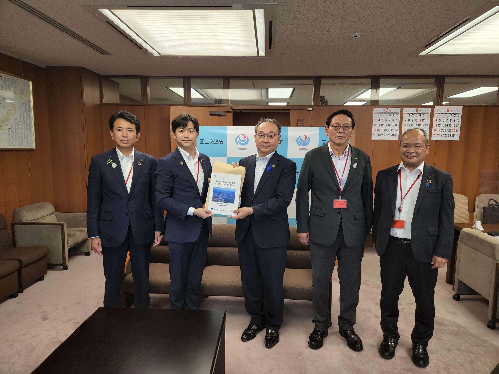 5名の男性が横一列に並び、中央に立つ吉岡幹夫技監と同盟会の男性が一緒に要望書を持っている集合写真