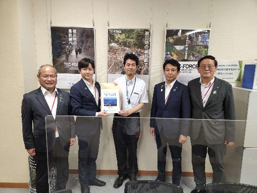 5名の男性が横一列に並び、中央に立つ岡村次郎に水管理・国土保全局長と同盟会の男性が一緒に要望書を持っている集合写真