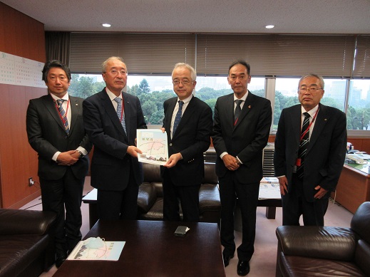 5名の男性が横一列に並び、中央に立つ野村正史国土交通審議官と同盟会の男性が一緒に要望書を持っている集合写真