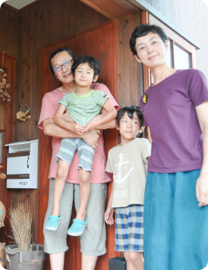 木目の自宅玄関の前で中年夫婦と小学校低学年くらいの男の子が2人うつっている家族写真