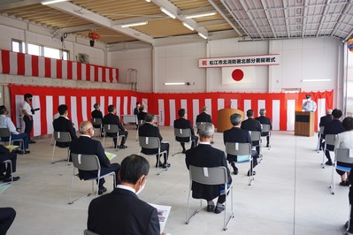 前方と左側の壁に紅白の幕が掛けられた会場の右奥の檀上に立つ男性の話を、間隔を開けて設置されたパイプ椅子に座っている関係者が聞いている北部分署開署式の様子の写真