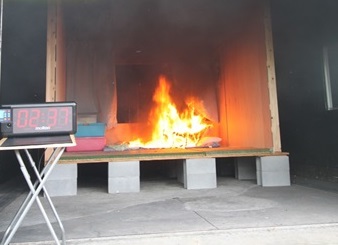 入口にタイマーが設置されその後ろの屋内で火災が起きている燃焼実験を行っている写真