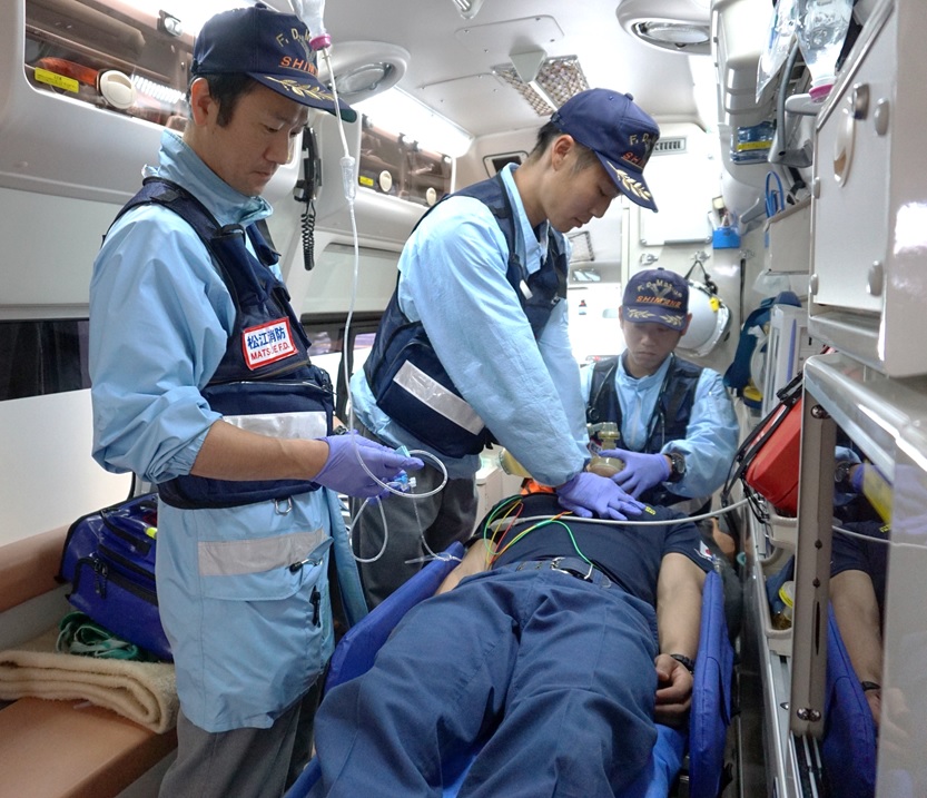救急車内酸素マスクや点滴、心臓マッサージを行っている3名の救急隊員の写真