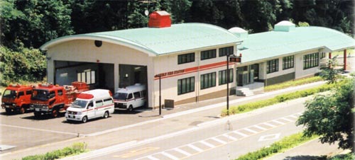 屋根が薄緑色の曲線造り、オフホワイト色の壁の西部分署に2台の救急車と2台の消防車が駐車している建物全景の写真