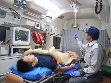救急車内でストレッチャーの乗った男性に救急隊が点滴処置をしている写真