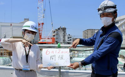 女性と工事関係者の男性が一緒に「6月12日（日曜）開催」と書かれたボードを指で差して持っている写真