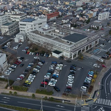 敷地内の前庁舎の周囲の駐車場に沢山の車が駐車している庁舎全景を上空から撮影した写真