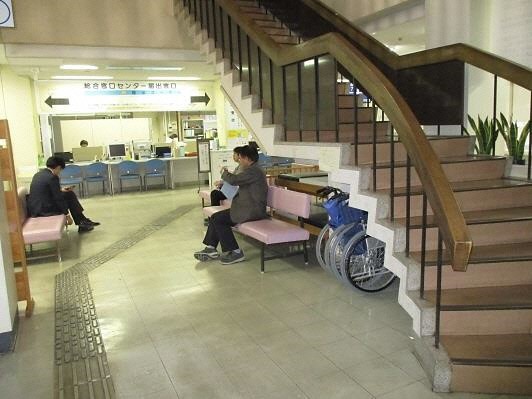 窓口の手前両側に設置されたピンク色の長椅子に来庁者が座り、右側に2階へ上がる階段がある市役所の待合スペースの写真