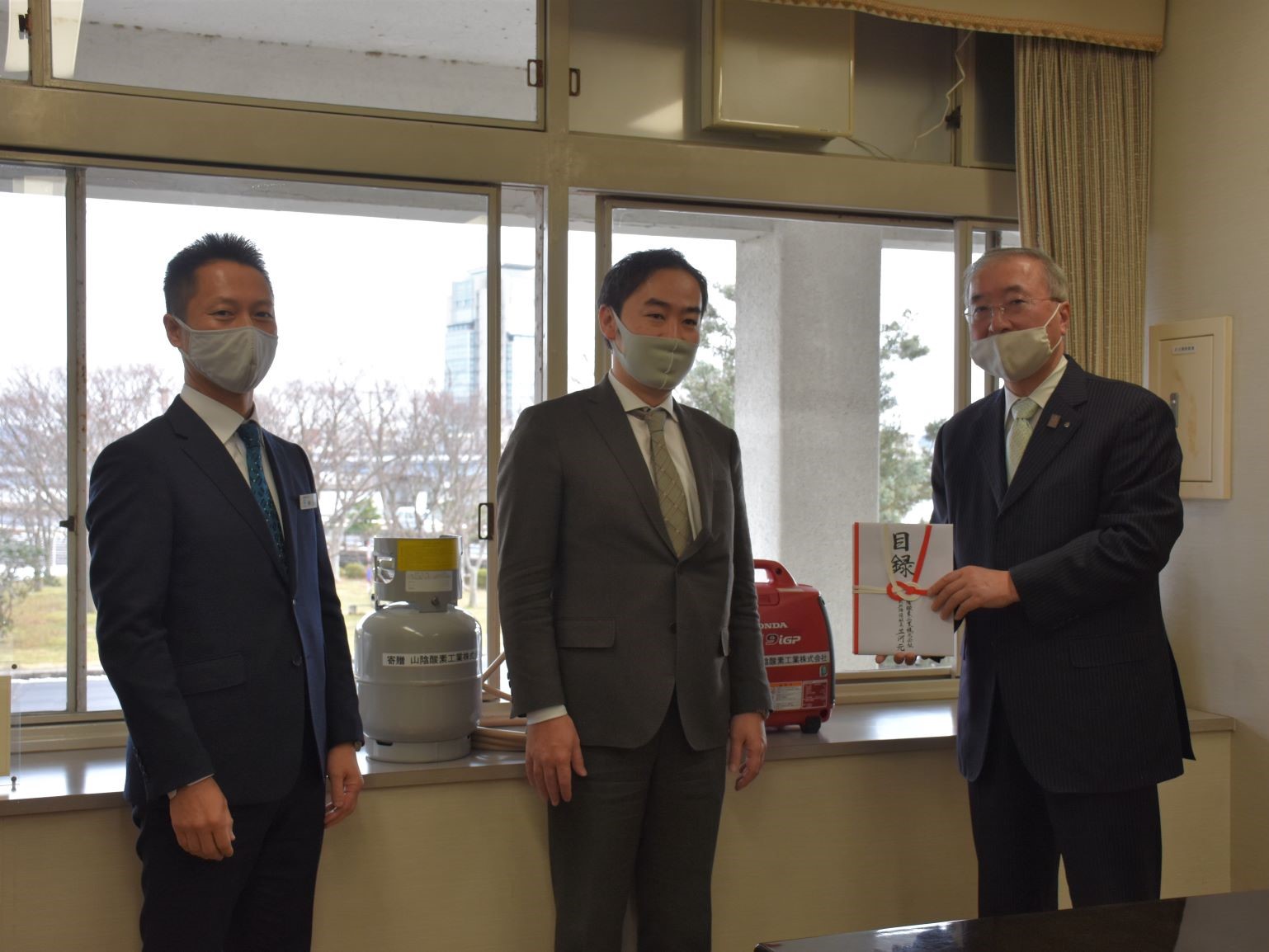 非常用発電機セットを背景に、目録を持っている松浦松江市長と、寄贈者の男性2人が並んでいる写真