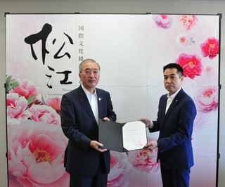 花が散りばめられたパネルを背景に、松浦松江市長と一緒に協定書を持っている出羽会長の写真
