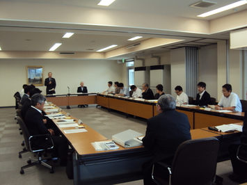 長方形に置かれた長机に出席者が座り奥の男性が立って話をしている様子の写真