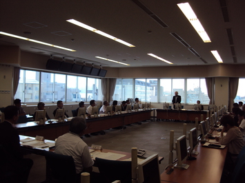 広い会議室で全員が内側を向く形で四角に机が置かれ奥の男性が立って発言する様子の写真