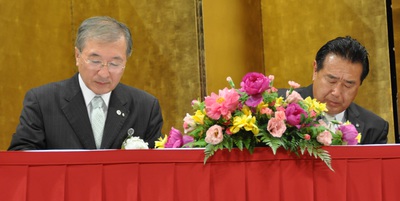 背後には金の屏風が立てられ、赤のテーブルクロスが敷かれ、お祝いの壇上花が置かれた席につき調印を行う鈴木町長と松浦市長の写真