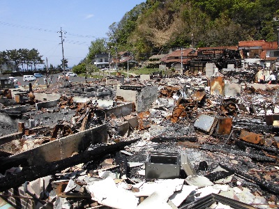 原型を留めないがれきの奥に焼損した建物のある被災地の写真