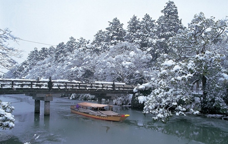 雪景色の松江城の敷地内の橋の下を遊覧船が渡っている写真