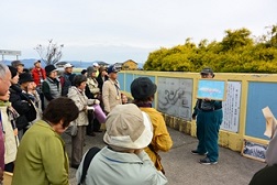 絵を見せながら説明をしている男性と男性の周囲に集まって話を聞いている参加者の写真