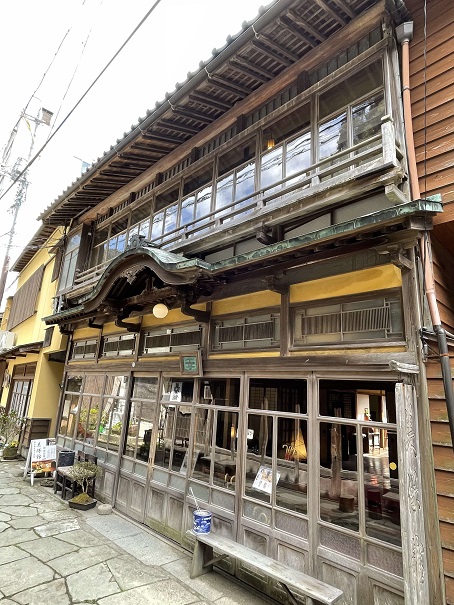 通り沿いにガラス窓、玄関の屋根に唐破風が施された木造造りの旅館美保館本館の建物外観の写真