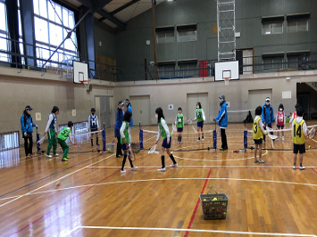 令和2年度の放課後子ども教室でのミニテニスの活動を写した写真、緑色と黄色と赤色と青色のビブスをつけた子供と青い服装の大人が計16人写っている