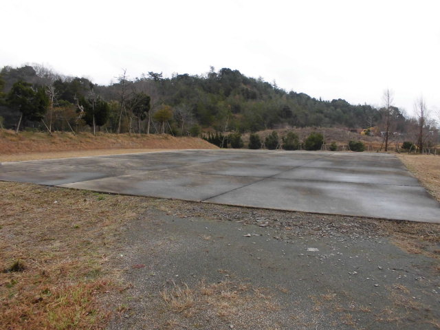 奥に山が見え、手前を樹木が生い茂る、コンクリートで平たく整地された広い土地の写真