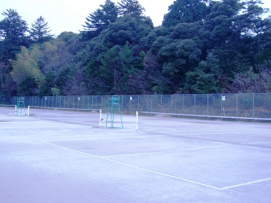 フェンスの奥に生い茂る森林を背景に、3面のテニスコートを斜め前から撮影した写真
