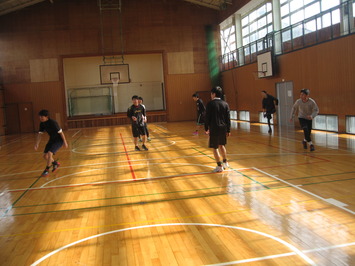 2階の窓から日が差し奥にバスケットゴールが見える体育館で、黒や灰色のスポーツウェアに身を纏った7人の男子が、ある一点に注目して身構えている様子の写真