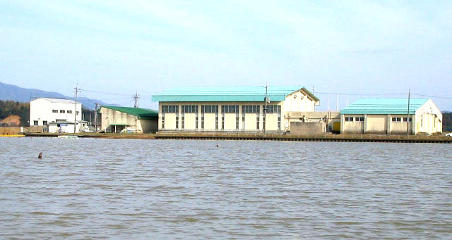 淡い水色の空を背景に、水色の屋根と白い壁を備えた大きな建物が水辺に立ち並ぶ施設群を、湖沼上から撮影した写真