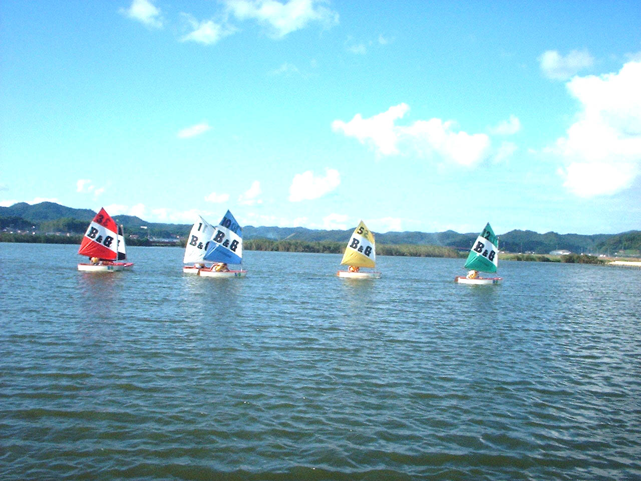 青い空と白い雲を背景に、西潟ノ内の湖沼上に浮かぶ、赤色、白色、青色、黄色、緑色の各ヨットを撮影した写真