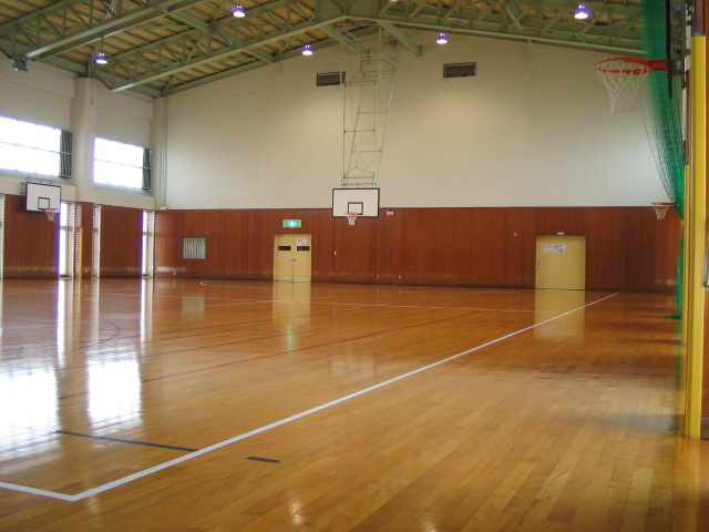上半分が白く下半分が茶色い壁で囲まれた体育館で、奥に縦横に設置されたバスケットゴールと、高い位置に設置された大きな窓から日が差している様子を、斜めから撮影した写真