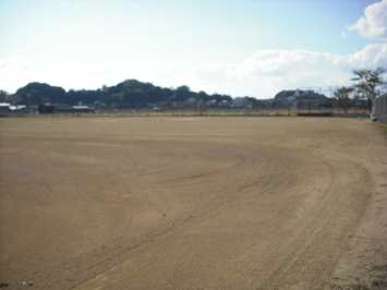 淡い水色の空を背景に、奥に緑の山が見え、その手前にある茶色の土で敷き詰められた広大な敷地を撮影した写真