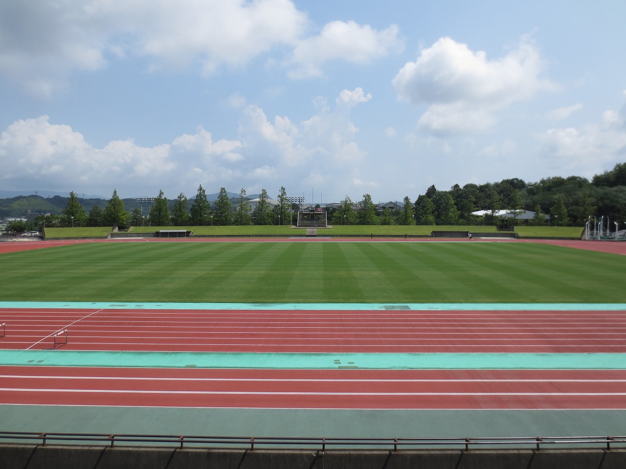 白い雲と青い空を背景に、中央に芝生の生えたグラウンドとその周囲を囲む赤い競走用トラックのある競技場を、真横から撮影した写真