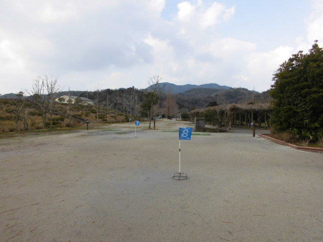 遠くに山が見え、周囲に枯れた木と白い文字で番号が書かれた青い旗が点在する、乾いた土がむき出しの地面のゴルフ場を撮影した写真