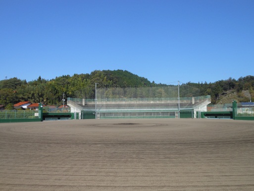 青空の下緑豊かな山を背景に、美保関総合運動公園野球場の均されたグラウンドとメインスタンドを写した写真